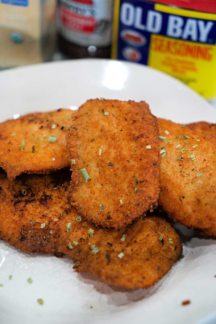Golden, crispy breaded chicken cutlets on a plate, garnished with herbs, with Old Bay seasoning and a side of fried tilapia for the perfect tilapia recipe in the background.