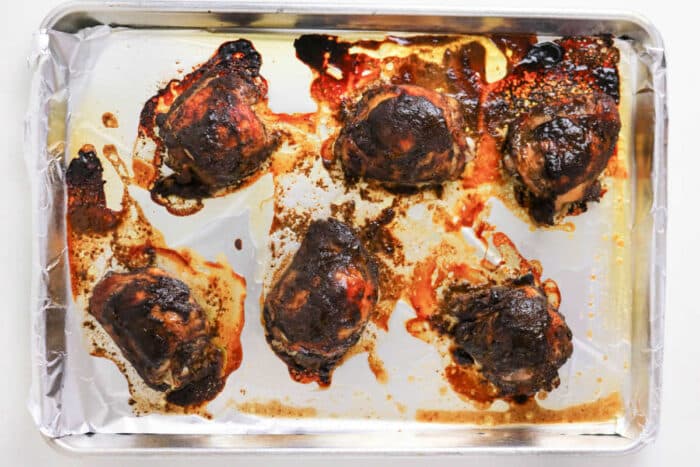 Six oven jerk chicken thighs on a foil-lined baking sheet, with browned and crispy skin and flavorful juices around them.