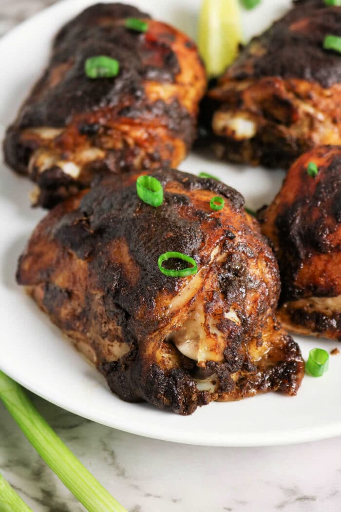 Four pieces of dark, grilled Jerk Chicken garnished with green onions, served on a white plate.