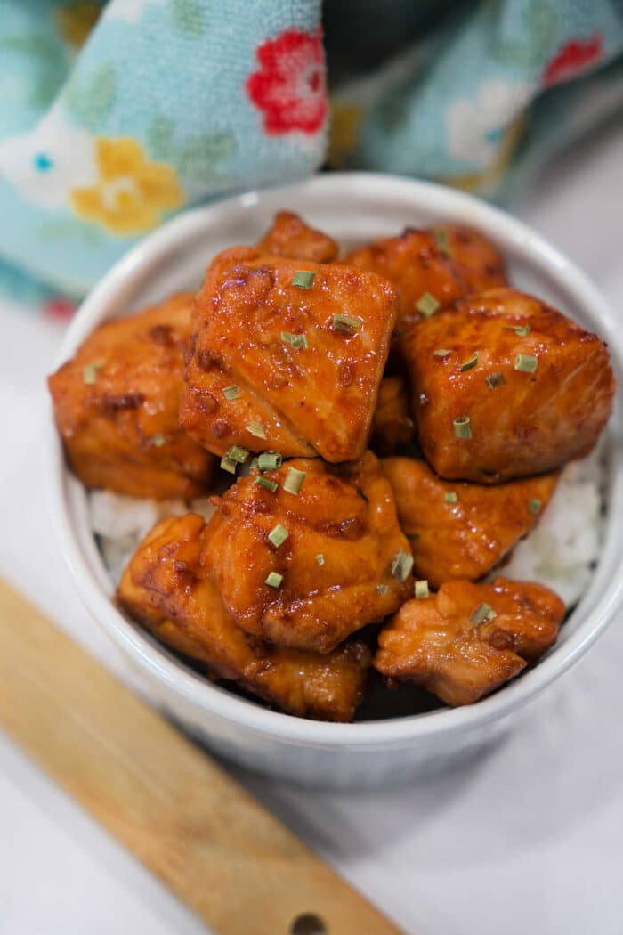 Bowl of Teriyaki Salmon Bites on white rice, garnished with herbs, with a patterned cloth in the background.