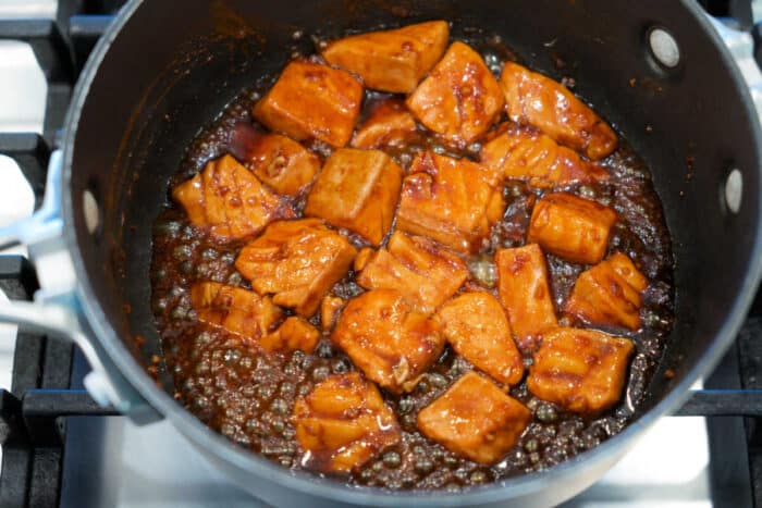 Chunks of Teriyaki Salmon Bites cooking in a dark, glossy sauce in a saucepan on a stovetop.