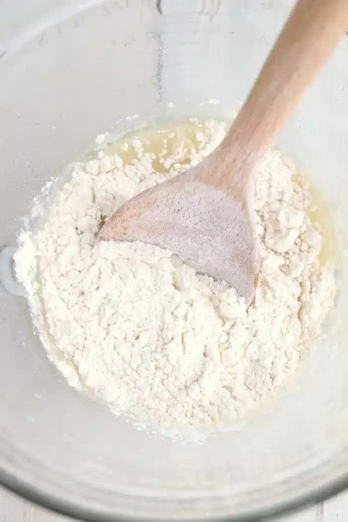 Wooden spoon mixing flour and liquid in a clear glass bowl, preparing dough for delicious air fryer bread.