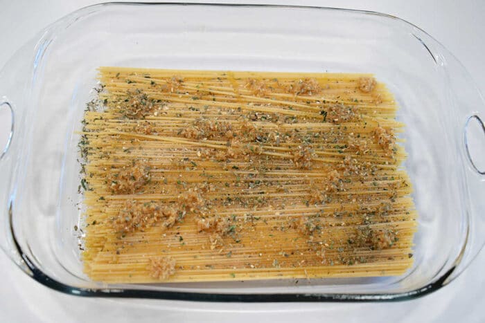 Uncooked spaghetti in a glass baking dish, topped with minced garlic and dried herbs, ready to be transformed into a classic baked spaghetti with meatballs.