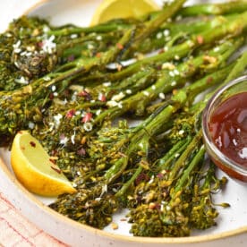 Oven roasted broccolini with lemon wedges and dipping sauce on a plate, sprinkled with sesame seeds and spices.