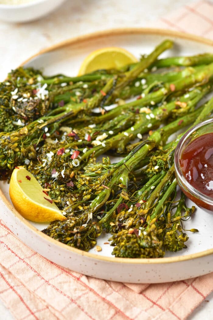 Oven roasted broccolini with lemon wedges and dipping sauce on a plate, sprinkled with sesame seeds and spices.