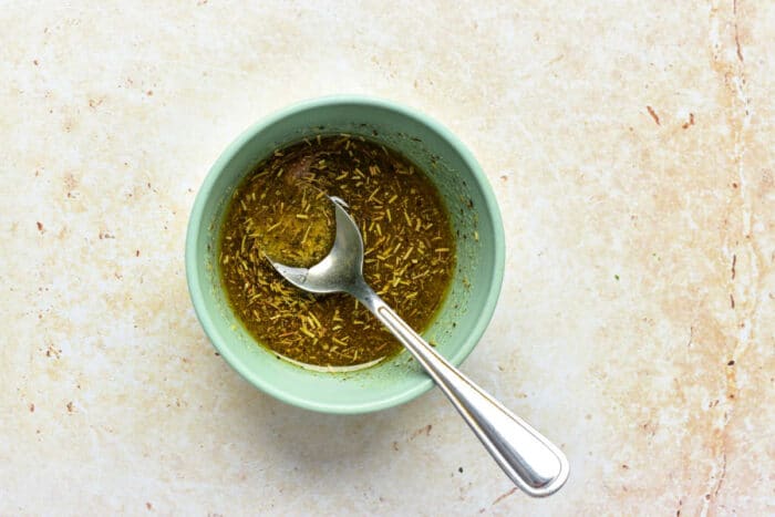 A small blue bowl with herb marinade and a spoon—a perfect accent for oven roasted broccolini—rests on a light textured surface.
