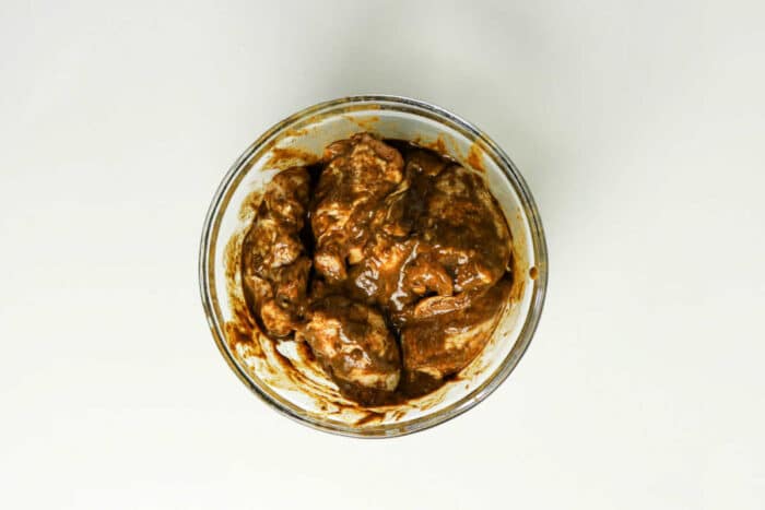Glass bowl with pieces of meat coated in a thick brown jerk chicken marinade, ready for the oven, placed on a white surface&mdash;a perfect start to your favorite jerk chicken recipe.
