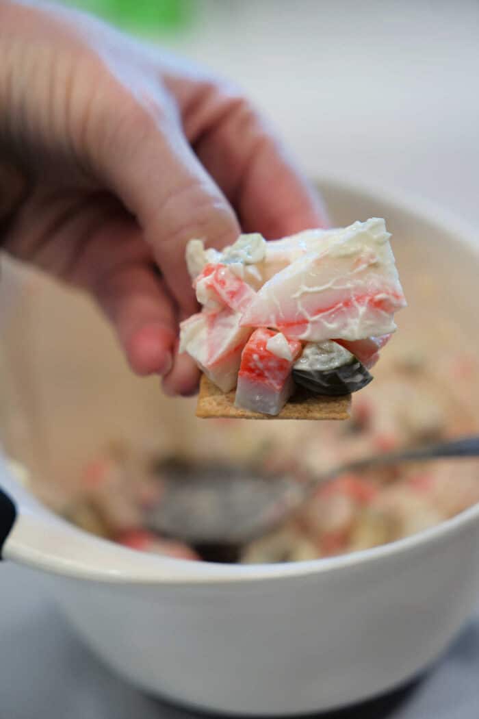 A hand holds a cracker topped with creamy crab salad above a white bowl filled with the seafood sandwich mixture.