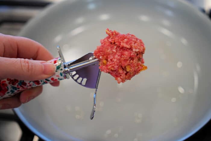Hand holding a scoop of raw ground meat over an empty frying pan on a stove, perfect for preparing baked spaghetti with meatballs.