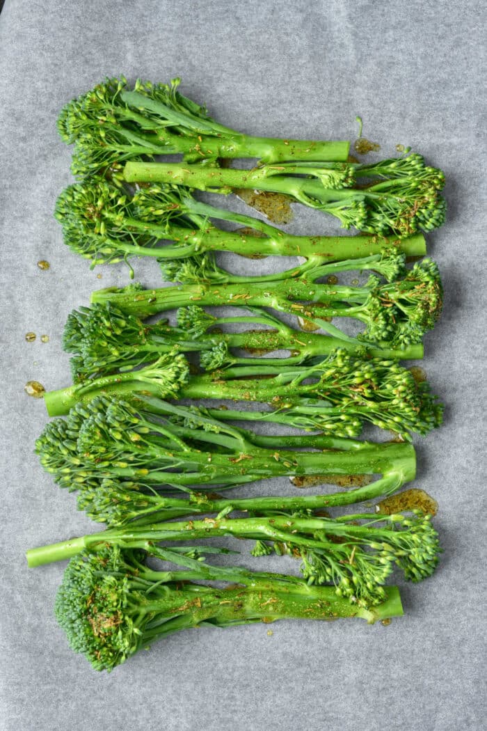 Fresh broccolini with seasoning drizzled on top, arranged on parchment paper, ready to be transformed into delicious oven roasted broccolini.