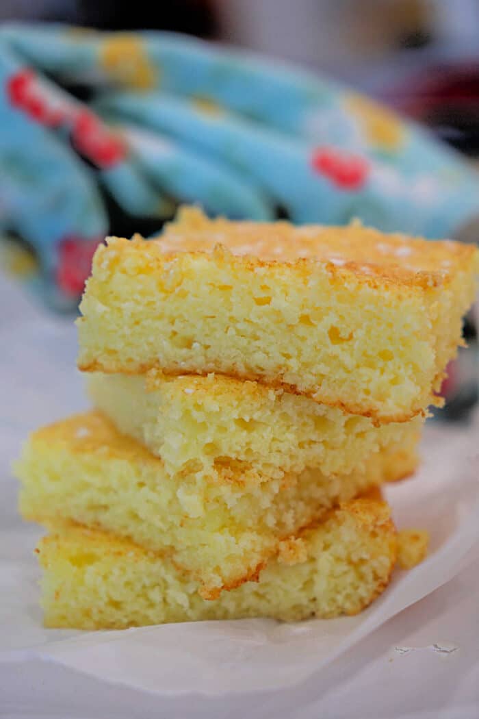 Three pieces of cornbread stacked on parchment paper, with a colorful cloth in the background—an inviting scene reminiscent of a fresh lemon angel food cake display.
