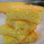 Three pieces of golden, crumbly cornbread stacked on a sheet of white parchment paper, reminiscent of the airy texture found in lemon angel food cake.
