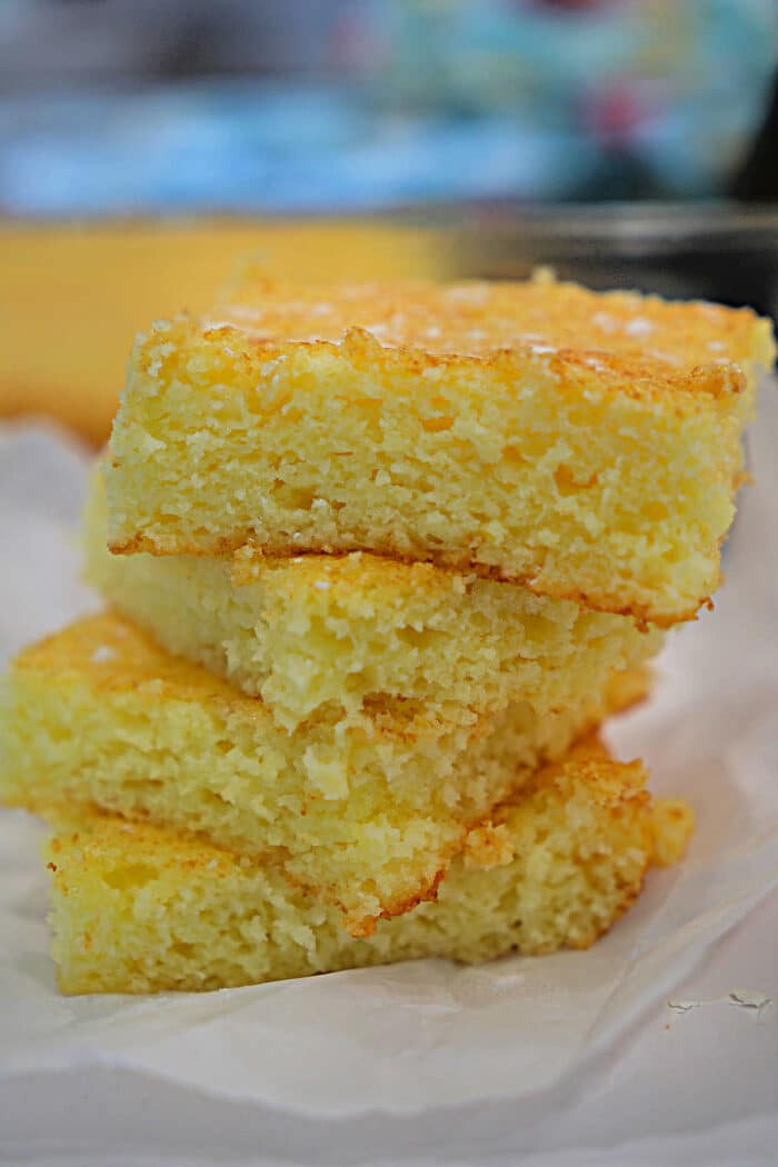 Three pieces of golden, crumbly cornbread stacked on a sheet of white parchment paper, reminiscent of the airy texture found in lemon angel food cake.