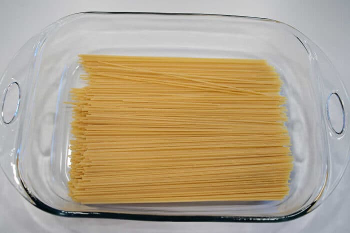 Uncooked spaghetti noodles arranged in a clear glass baking dish on a white surface, ready to be transformed into delicious baked spaghetti with meatballs.