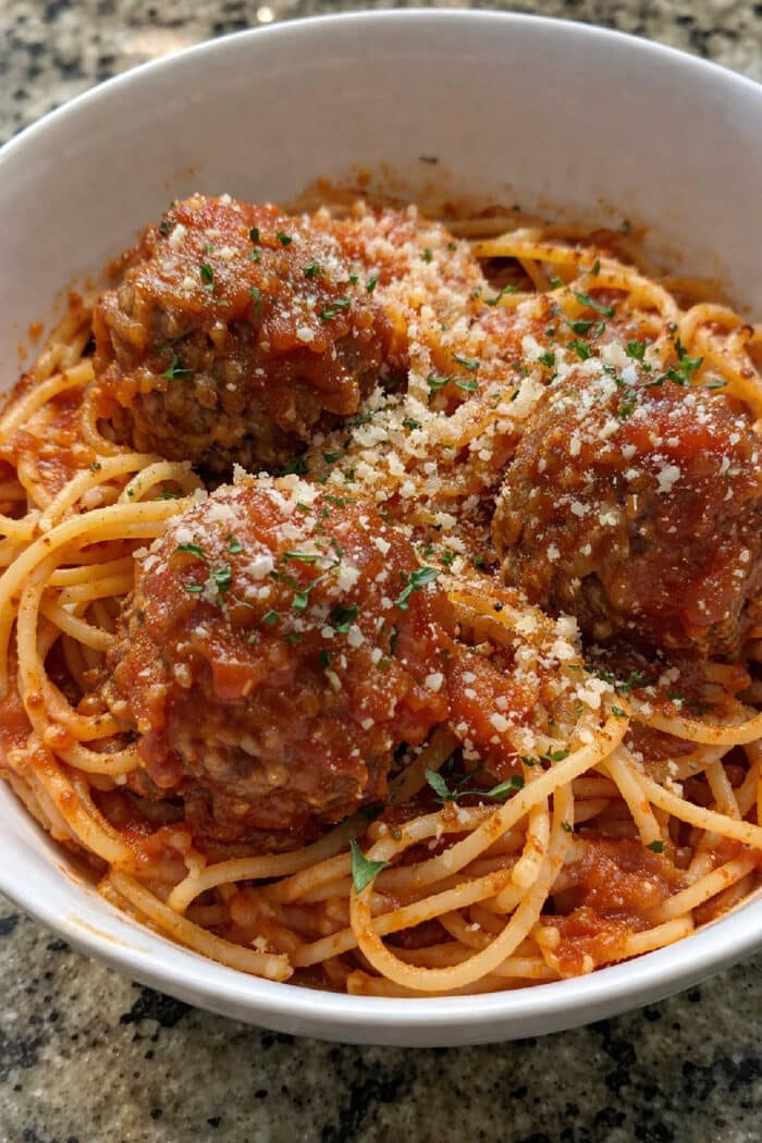 Baked spaghetti with meatballs topped with tomato sauce, grated cheese, and herbs, served in a white bowl on a granite counter.