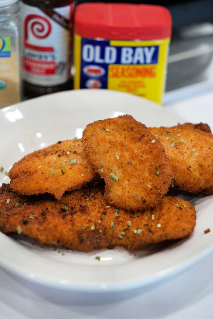 A plate of golden, breaded fried chicken cutlets, garnished with herbs, with seasoning containers behind—perfect for those who love a great Fried Tilapia or classic chicken dish.