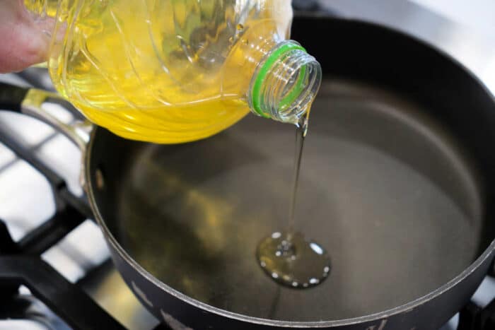 Vegetable oil being poured from a plastic bottle into a black frying pan on a stovetop, ready for preparing a delicious fried tilapia recipe.