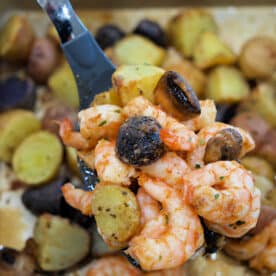 A spoon holds shrimp and potatoes with mushrooms above a tray filled with roasted shrimp and potatoes, plus savory mushrooms.