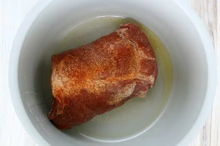 Raw, seasoned meat sits in oil at the bottom of a white slow cooker, ready to transform into tender ninja foodi pulled pork.