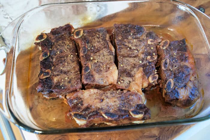 Five cooked beef short ribs in a glass baking dish, seasoned and ready to serve.