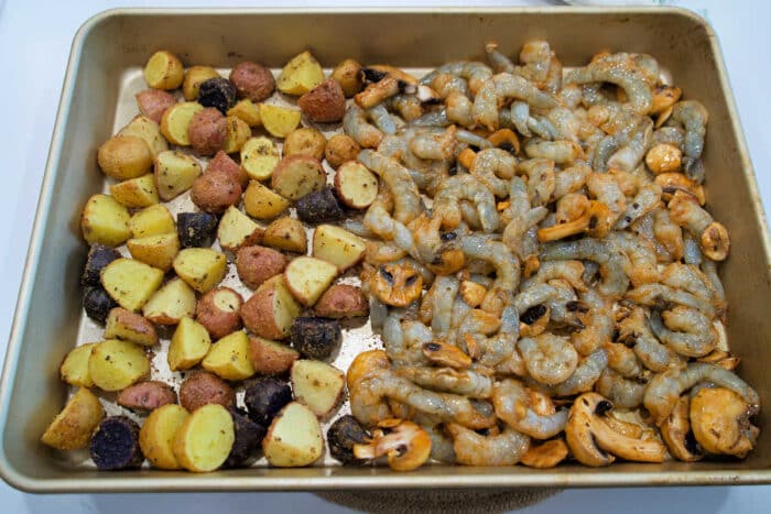 Sheet pan with seasoned baby potatoes on the left and raw shrimp and mushrooms on the right, this shrimp and potatoes combo is ready to bake.