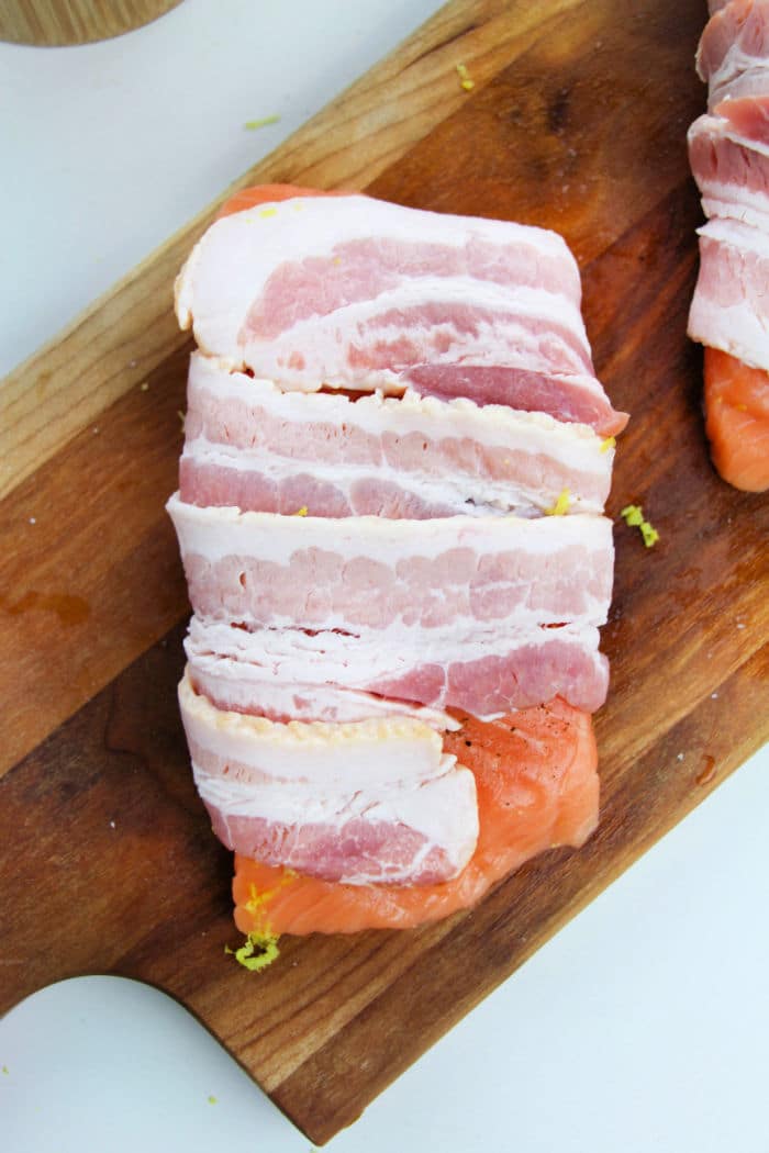 Bacon Wrapped Salmon features raw salmon fillet wrapped in strips of uncooked bacon, arranged on a wooden cutting board.
