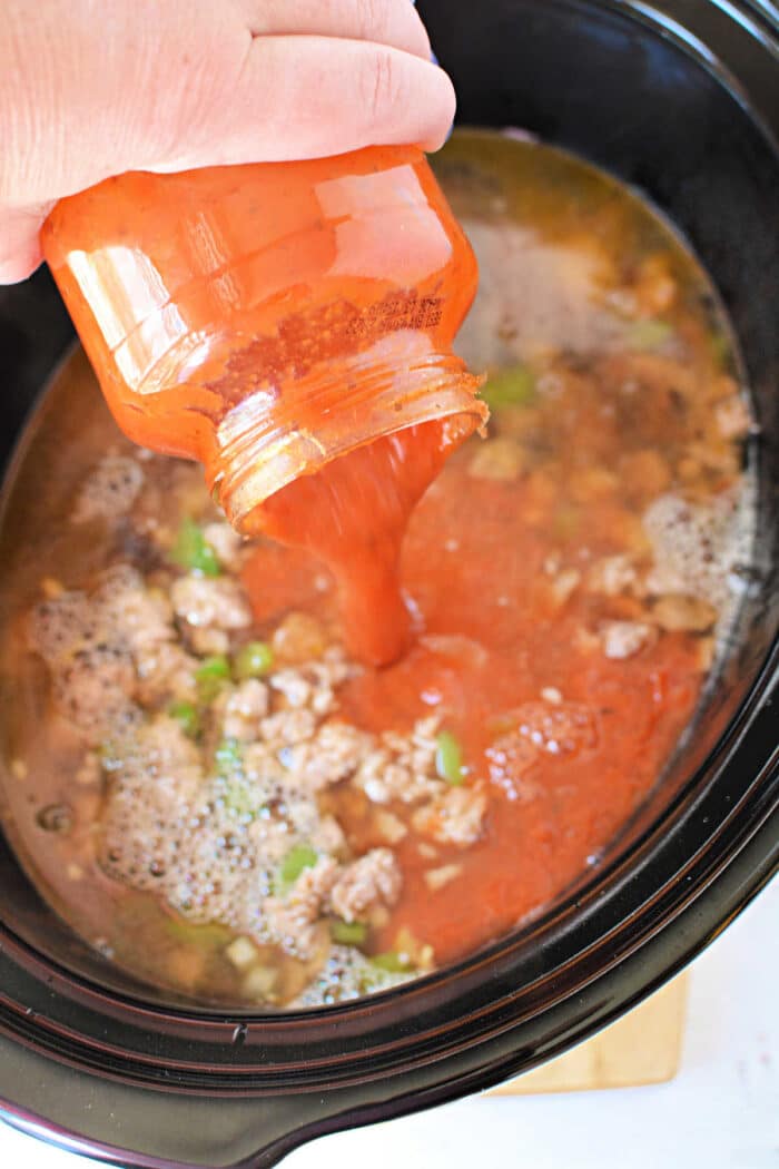 A hand pours tomato sauce from a jar into a Crockpot filled with ground meat and vegetables, perfect for making hearty Lasagna Soup.
