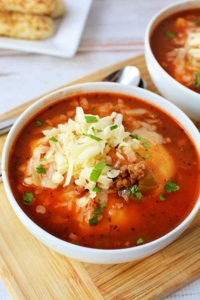 A bowl of hearty Slow Cooker Lasagna Soup topped with shredded cheese and chopped herbs sits on a wooden board.