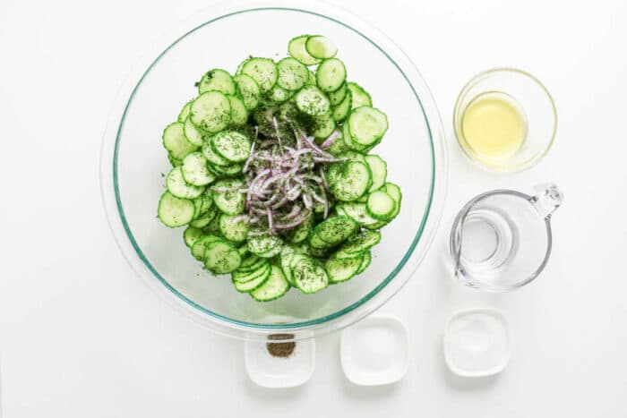 Glass bowl with sliced cucumbers, onions, and fresh dill, creating a crisp cucumber salad, surrounded by vinegar, water, salt, sugar, and pepper.