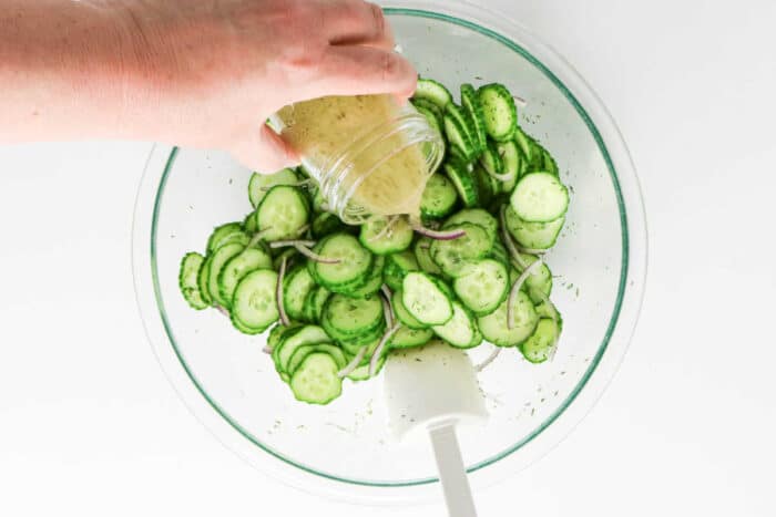 A hand pours dressing over sliced cucumber and onion in a glass bowl with a white spatula, creating a fresh salad perfect for adding dill.