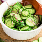 Bowl of cucumber salad with crisp red onion slices and fresh dill, all mixed together with a wooden spoon.