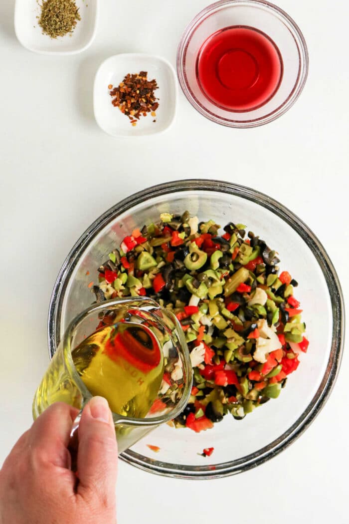 A hand pours olive oil into a bowl of chopped vegetables for a classic Olive Salad, with spices and red vinegar nearby&mdash;perfect for an authentic Muffuletta recipe.