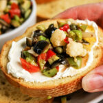 A hand holds a slice of bread topped with cream cheese and a vibrant Olive Salad, featuring chopped olives, cauliflower, and peppers—a perfect bite inspired by the classic Muffuletta recipe.