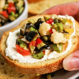 A hand holds a slice of bread topped with cream cheese and a vibrant Olive Salad, featuring chopped olives, cauliflower, and peppers&mdash;a perfect bite inspired by the classic Muffuletta recipe.
