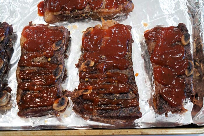 Three pieces of barbecued Flanken Short Ribs topped with sauce on a foil-lined baking sheet.