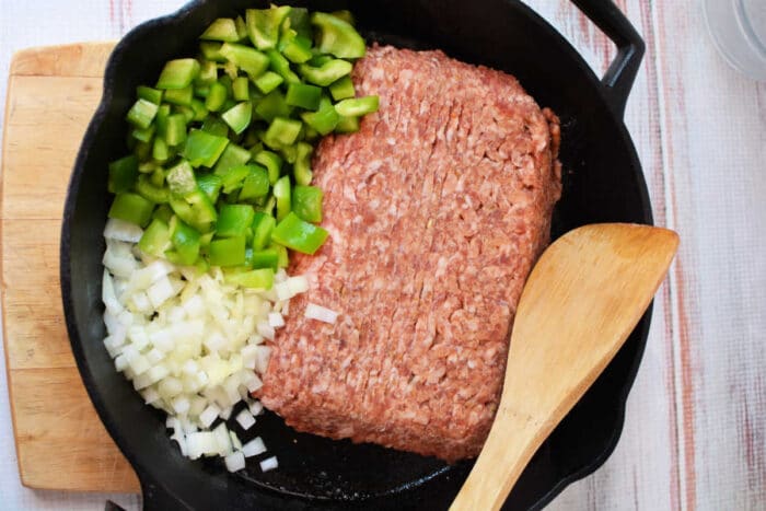 Ground meat, chopped green bell pepper, and chopped onion in a cast iron skillet with a wooden spatula&mdash;perfect for starting your Slow Cooker Lasagna Soup.
