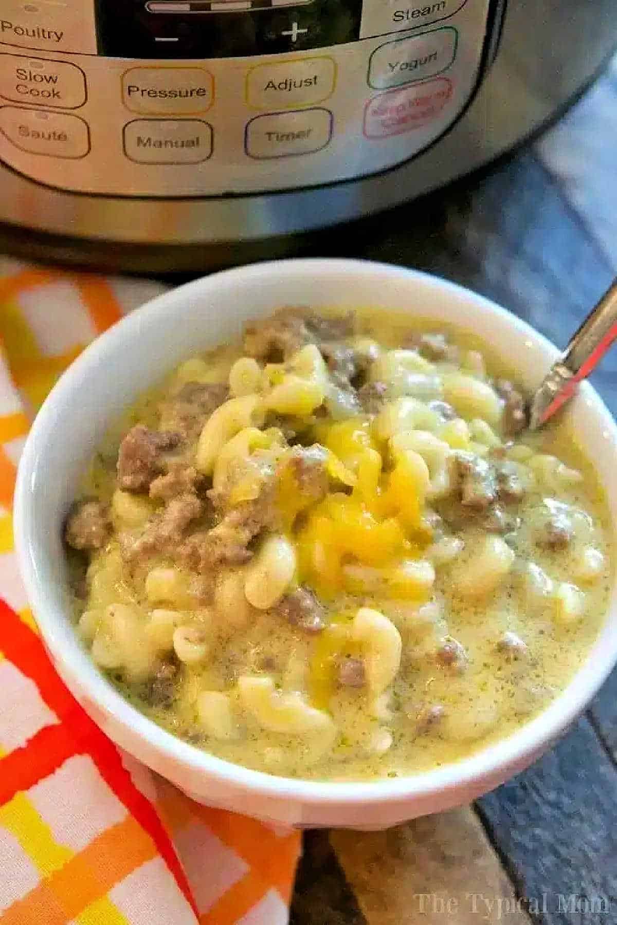 This Instant Pot cheeseburger macaroni recipe will take you back to your childhood! Just 10 minutes in your pressure cooker makes this cheesy goulash hamburger helper style pasta dinner that your whole family will love. It's one of our favorite Instant Pot ground beef recipes. #instantpot #pressurecooker #cheeseburger #macaroni #goulash #hamburgerhelper #groundbeef #instantpotrecipes #thetypicalmom Bowl of Instant Pot cheeseburger macaroni with a spoon, next to an Instant Pot and a colorful napkin.