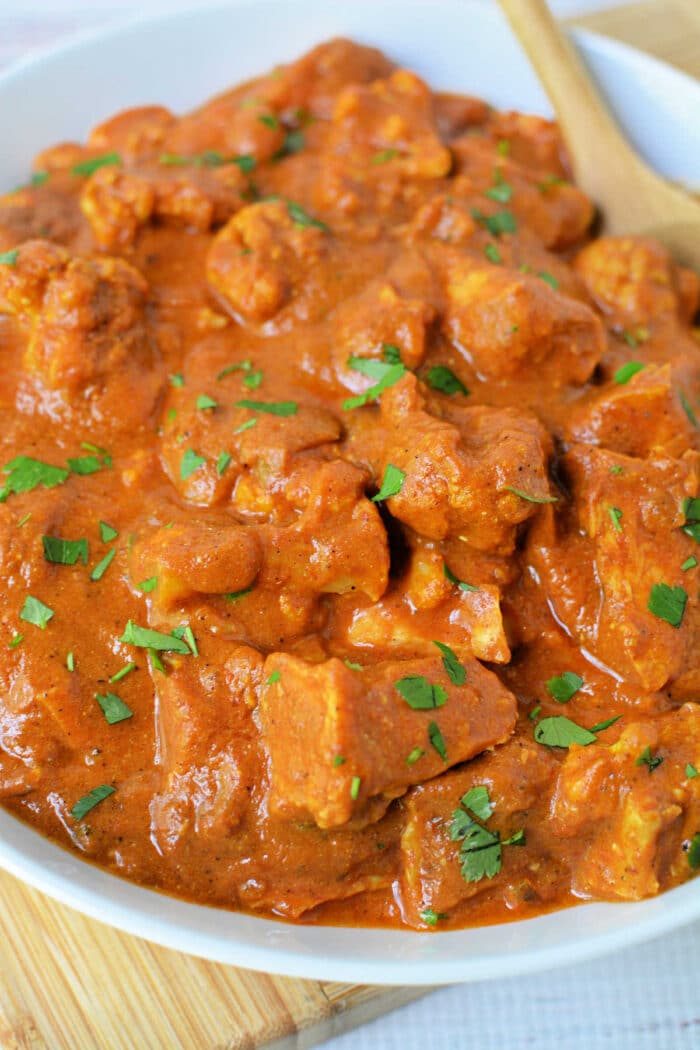 A close-up of slow cooker chicken tikka masala, reminiscent of butter chicken, garnished with chopped cilantro in a white bowl with a wooden spoon.