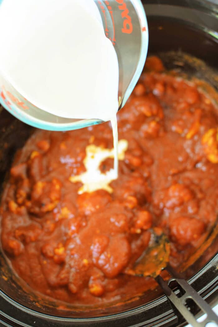 Milk is being poured from a measuring cup into a slow cooker filled with a tomato-based sauce, perfect for making Slow Cooker Chicken Makhani.