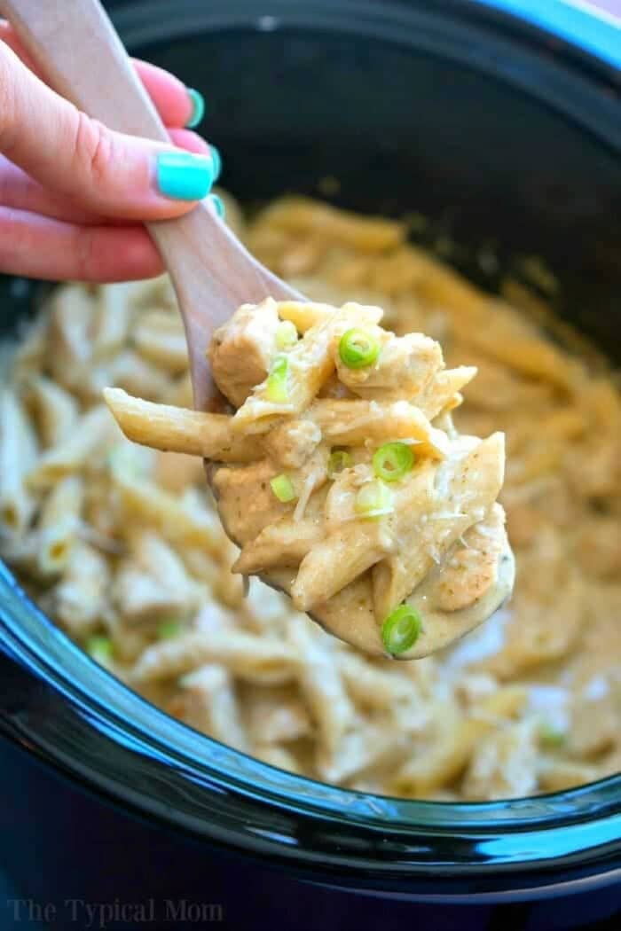 This easy crockpot pesto chicken alfredo casserole is a hit in my house! Cheesy noodles with moist pieces of pesto chicken together makes a complete meal. A wooden spoon holds creamy pasta with green onions over a crockpot pesto chicken alfredo casserole, brimming with rich flavors.