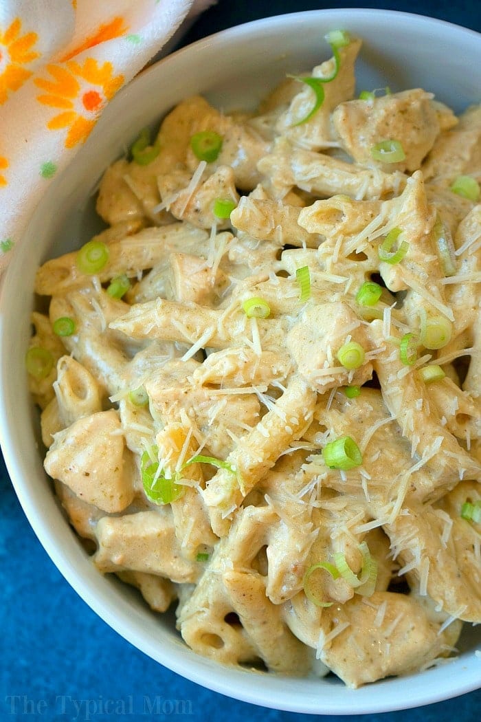 This easy crockpot pesto chicken alfredo casserole is a hit in my house! Cheesy noodles with moist pieces of pesto chicken together makes a complete meal. Bowl of creamy pasta with grated cheese and sliced green onions, next to a floral-patterned napkin—reminiscent of a hearty crockpot pesto chicken alfredo casserole.