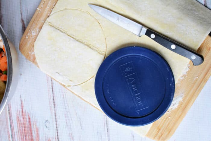 Round blue lid and knife on pastry dough with a circular shape cut out, ready for your homemade KFC Chicken Pot Pie Recipe, all set on a wooden board.