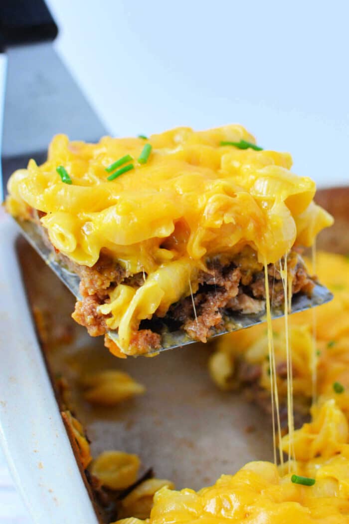 A spatula lifts a cheesy meatloaf casserole from a baking dish, topped with melted cheese and chives, blending classic casserole comfort with the creamy goodness of mac and cheese.