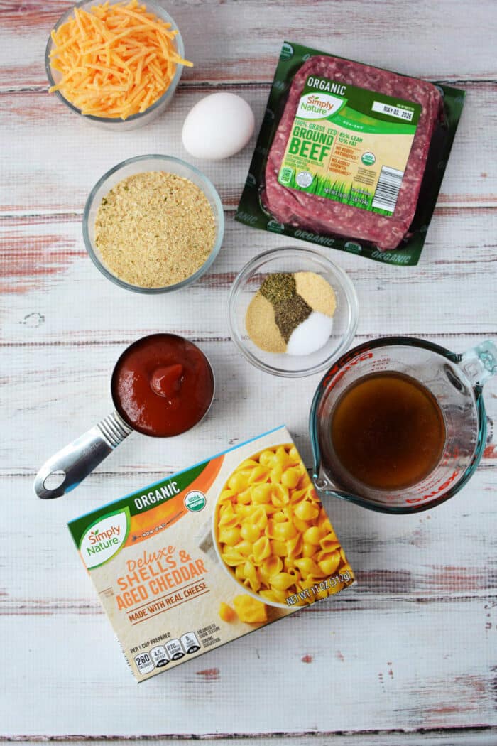 Ingredients for a cheesy casserole, blending pasta, ground beef, cheese, egg, broth, sauce, and seasonings—like a tasty mash-up of cheeseburger and classic mac and cheese.