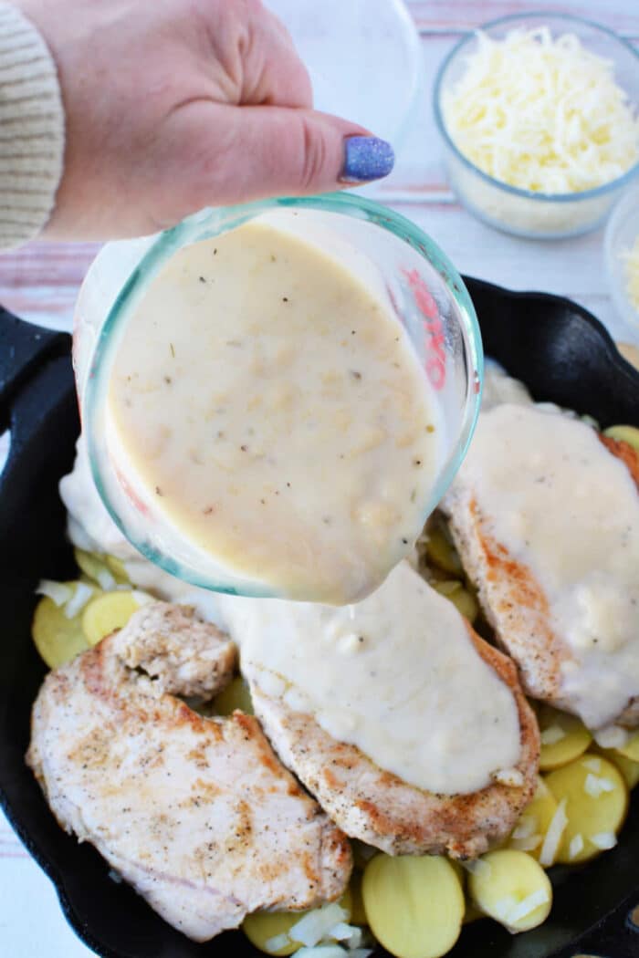 A hand pours creamy sauce over seasoned oven baked pork chops and sliced potatoes in a skillet.