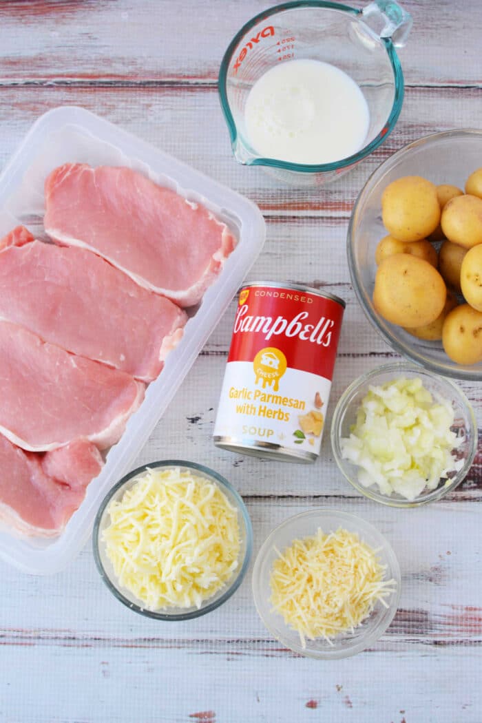 Raw pork chops with potatoes, chopped onions, shredded and grated cheese, milk, and a soup can are arranged on a table—perfect ingredients for delicious oven baked pork chops.