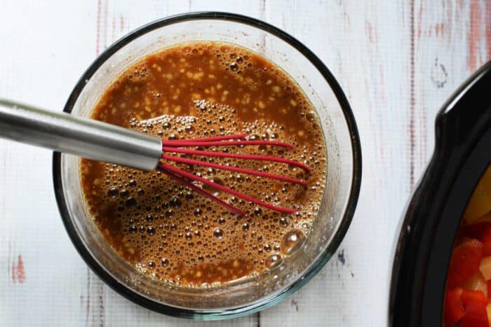A metal whisk with a red handle mixes a brown sauce—perfect for slow cooker chicken—in a glass bowl on a white wooden surface.