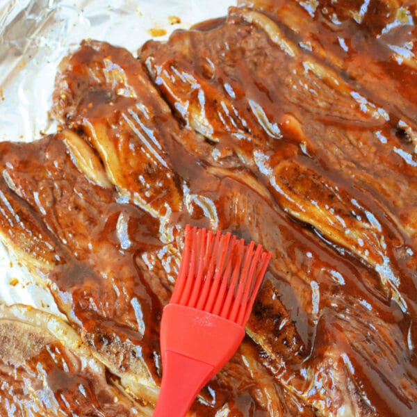 A red brush spreads barbecue sauce over beef riblets on a foil-lined baking sheet, making this the perfect beef riblets recipe for your next cookout.