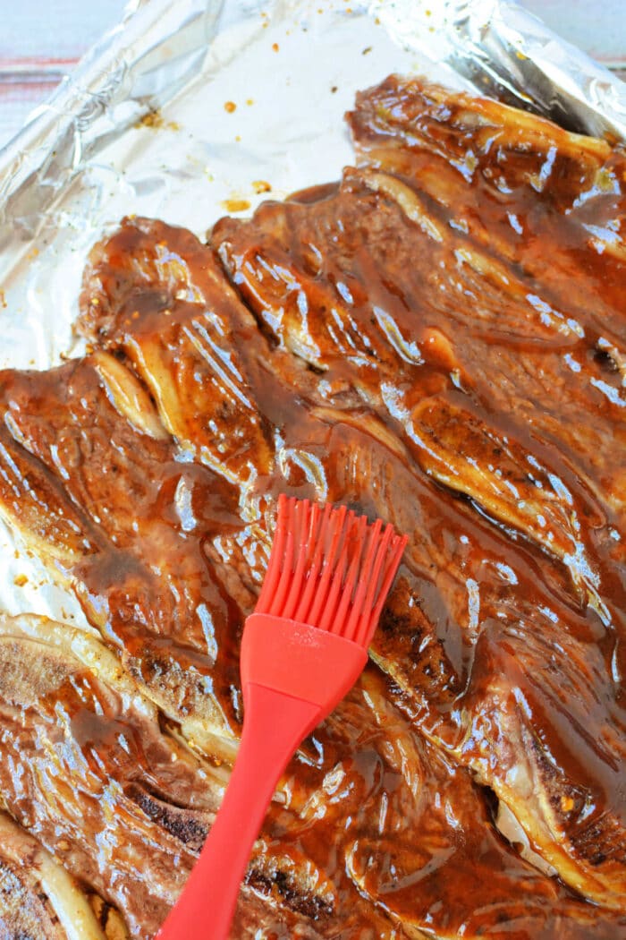 A red brush spreads barbecue sauce over beef riblets on a foil-lined baking sheet, making this the perfect beef riblets recipe for your next cookout.
