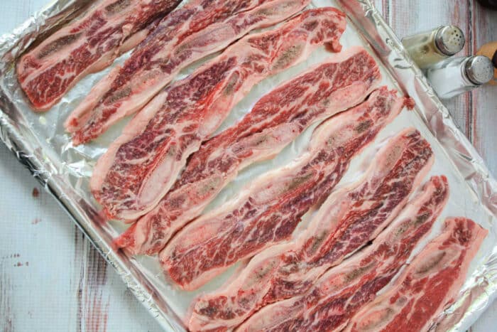 Raw marinated beef riblets arranged on a foil-lined baking sheet, with salt and pepper shakers nearby—perfect for trying out your favorite Beef Riblets Recipe.